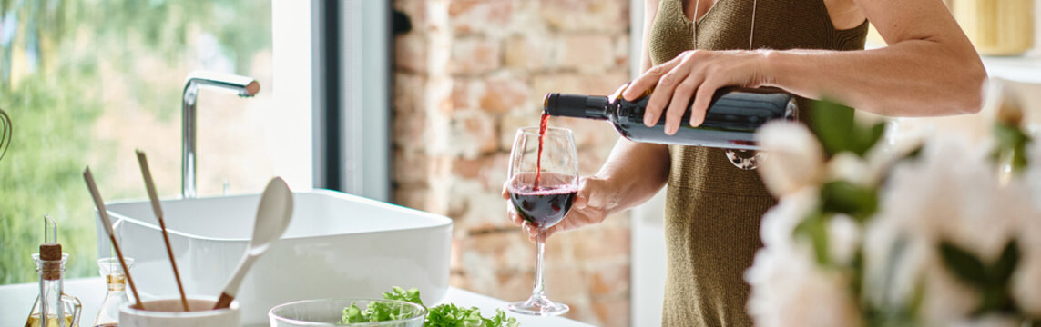 cropped view of woman pouring red wine into glass in modern kitchen at home, horizontal banner - Powered by Adobe