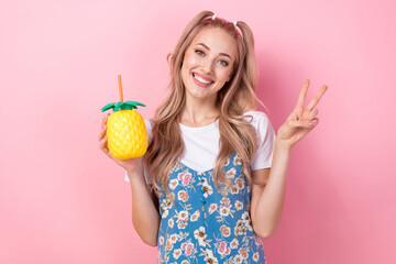 Portrait of pretty positive girl hold pineapple juice cup demonstrate v-sign isolated on pink color background