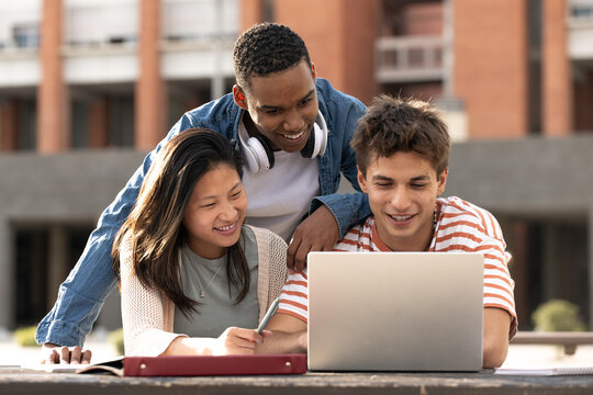 Diverse group of students working together with a laptop in campus.Young confident friends studying outside using computer and notes.