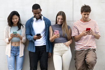 Diverse group of young people texting leaning against a wall. Happy group of friends using their...
