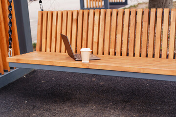 A cup of hot coffee and a gray laptop on a wooden bench. Online Work and Study Concept