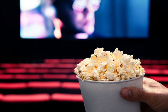 Movies and popcorn. Man holding pop corn box at cinema. Action, thriller or scifi entertainment on screen. Red seats in dark theater. Salty snack in bucket. Spectator pov. Film premiere.