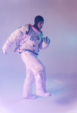 Male ethnic astronaut in spacesuit and backpack with dark hood marching against purple background