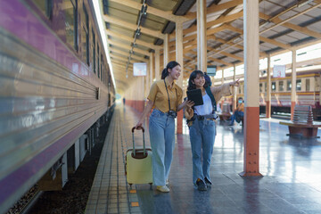 Two women are happy while traveling at the train station. tour concept. Two Asian women's best friends travel pointing fingers at a train timetable at a railway station.