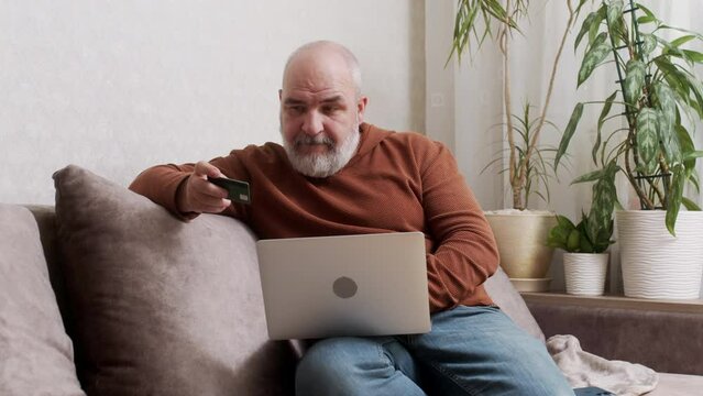 Remote Business. An Elderly Gentleman Using A Laptop And Talking On A Cell Phone Sitting On The Couch Of The House, An Elderly Man Working Online On A Computer And Having A Phone Conversation.