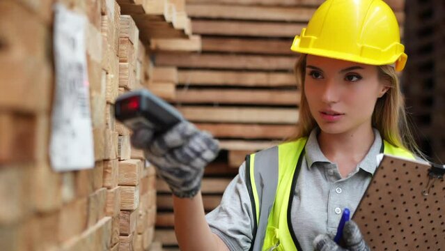 Warehouse Inventory Check: A focused female worker wearing a yellow hard hat and safety vest meticulously scans a barcode on a stack of wooden planks, clipboard in hand.