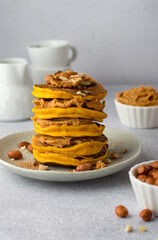 A stack of orange pumpkin pancakes, sprinkled with nuts, peanuts, cinnamon, peanut butter, white ceramic dish. An example of serving breakfast..