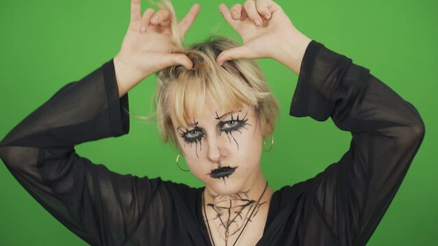 Portrait of a goth girl in black clothes and special makeup on a green background. She looks at the camera and makes horns on her head with her hands. Chroma key.