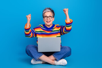 Full size portrait of delighted funky lady sit floor use netbook raise fists success isolated on blue color background