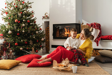  Young woman and her little son are having fun beside a glowing fireplace in a cozy living room...