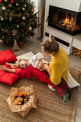  Young woman and her little son are having fun beside a glowing fireplace in a cozy living room adorned with a Christmas tree and festive decorations