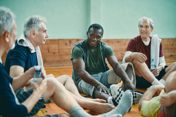 Diverse group of male senior friends sitting on the floor of a gym after playing basketball