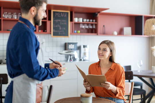 Friendly european male waiter writing down an order from young lady in coffee shop, woman holding menu and smiling to man
