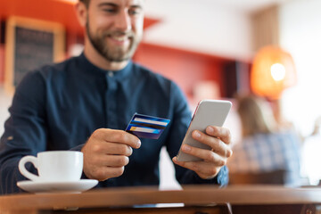 Handsome smiling european man with credit card and smartphone at cafe, drinking coffee and shopping online, purchasing in internet, focus on hands