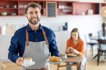 Friendly male waiter holding cup of coffee and plate with pastry, serving order for female customer, looking and smiling at camera in cafe