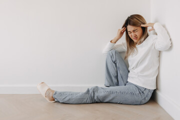 Obraz premium Asian Thai woman got headache with migraine, thinking too much, have no idea, tired sitting on floor and leaning the wall in apartment room winter.