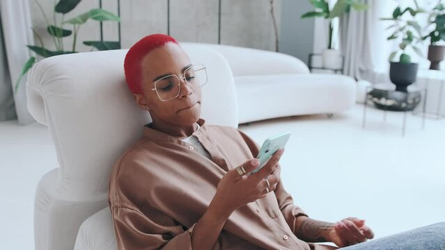 Young dissatisfied irritated African American woman holds mobile phone and begins to nervously swear and wave hand after learning about cancellation of weekend sits in chair in living room.