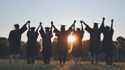 College graduates in robes holding hands at sunset.