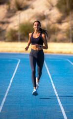 Beautiful slim young tanned runner girl, dressed in tight sportswear, smiling happily and enjoying running energetically on a blue track, approaching and looking at the camera.