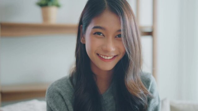 Confident smiling young adult asian woman looking at camera while sitting at home, Happy beautiful lady pretty face dental smile posing alone indoors, Slow motion close up view portrait