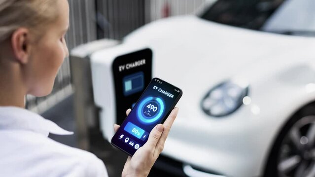 Businesswoman holding smartphone display battery status interface by smart EV mobile application while eco-friendly EV car recharging electricity from charging station in car park. Peruse