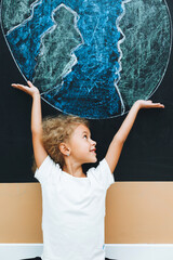 A cute little girl stands with her back and draws a planet on the board. the child holds his hands like a house above the planet.