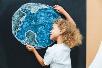 A cute little girl stands with her back and draws a planet on the board. the child holds his hands like a house above the planet.