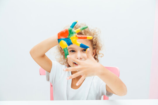 little blonde girl shows her hands painted with multi-colored paint on a white background.