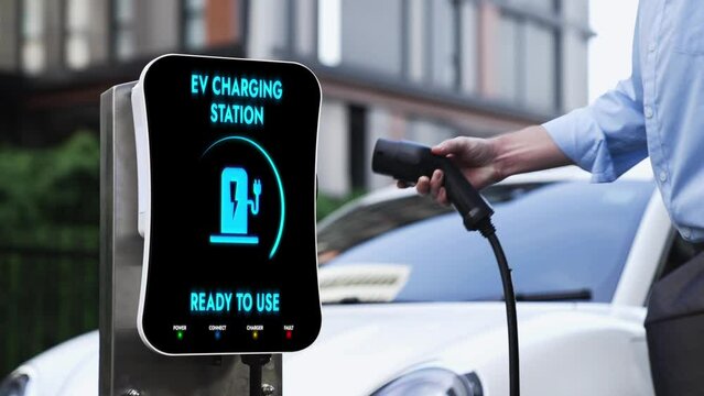 Businessman pull out EV charger from charging station to recharge electric car at city residential building. Modern urban lifestyle and eco-friendly electric car utilization to reduce CO2.Peruse