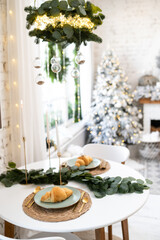 Decorated table in New Year's style. On the table is a plate with a croissant and candles against a background of a white Christmas tree and Christmas lights.