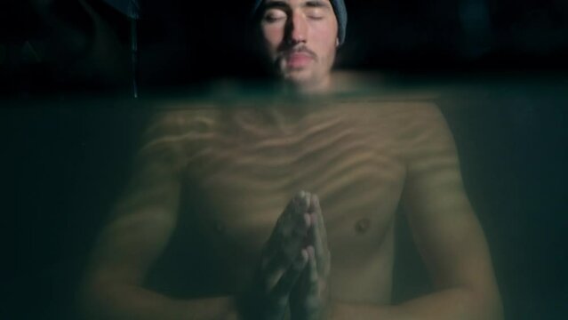 focused meditating man with closed eyes doing cold bath in the lake at night shaking hands under water revealing face on the surface