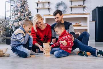 Happy young family with kids in woolen jumpers spending winter holidays at home playing with wooden blocks on warm floor in cozy, festively decorated living room with christmas tree - 661197561
