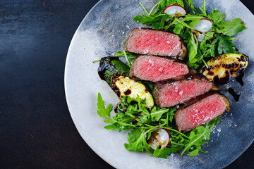 Barbecue dry aged angus roast beef steak with grilled zucchini slices and rocket lettuce served as top view in a Nordic design plate with copy space left