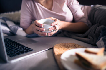 Close up of a young woman having snacks and coffee while using a laptop in bed