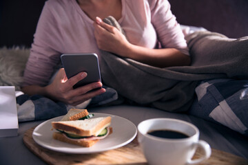 Fototapeta na wymiar Close up of a young woman using a smartphone having breakfast at bed