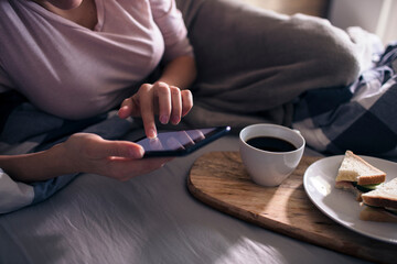 Fototapeta na wymiar Close up of a young woman using a smartphone having breakfast at bed