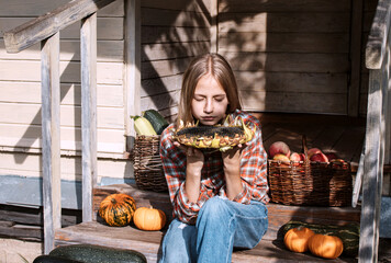 Girl is sitting on steps of old village house holding large sunflower in hands. Ripe pumpkins lie nearby and there are baskets filled with harvested crops: apples and zucchini. Harvest. Autumn. Fall