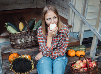 Girl is sitting on steps of an old village house with her face exposed to warm sunbeams and eating apple. Ripe pumpkins, baskets with apples and zucchini are nearby. Harvest. Fall. Thanksgiving day