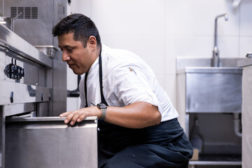 Latin chef opening the metal door of the fridge at the bottom of the kitchen