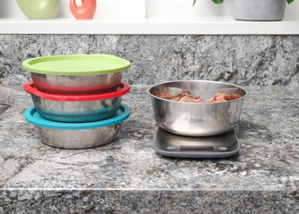 Raw food meal prep for dog in kitchen. Multiple pet food dishes and with scale. Measured ground chicken meat in portion and storing in stainless steel bowls with lids. Selective focus.