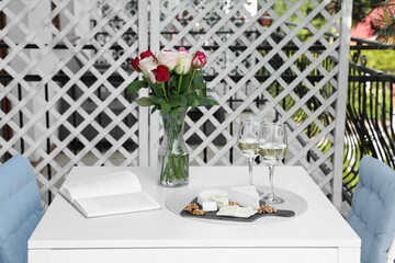 Vase with roses, open book, glasses of wine and snacks on white table at balcony
