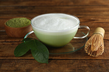 Cup of fresh matcha latte, bamboo whisk and green leaves on wooden table, closeup