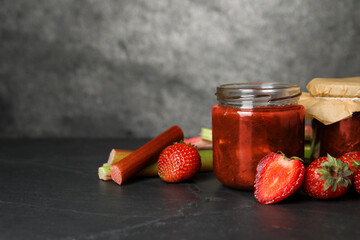 Jars of tasty rhubarb jam, fresh stems and strawberries on dark textured table. Space for text