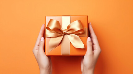 A woman's hands hold an orange gift box with a gold bow and ribbons on a colorful background - obrazy, fototapety, plakaty