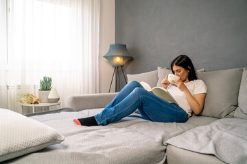 Pretty dark hair woman resting in cozy living room drinking tea and reading a book 