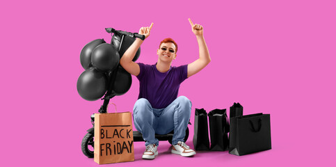 Happy young man with kick scooter, balloons and shopping bags pointing at something on magenta background. Black Friday sale
