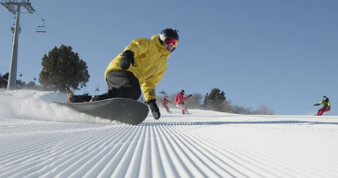 Group of people snowboarding on ski resort slope, snow spraying into the camera. Snowboarders carving on the fresh groomed corduroy slope during vacation on a sunny winter day