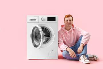 Young man sitting near washing machine on pink background