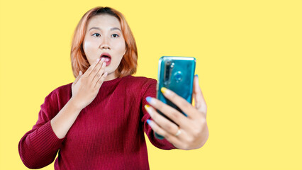 Shocked Asian woman looking at her mobile phone isolated on yellow background