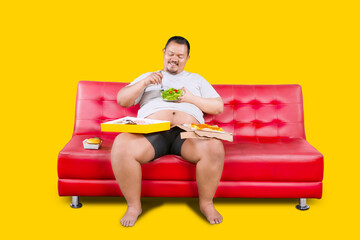 Overweight asian guy eating healthy food over unhealthy food isolated on yellow background